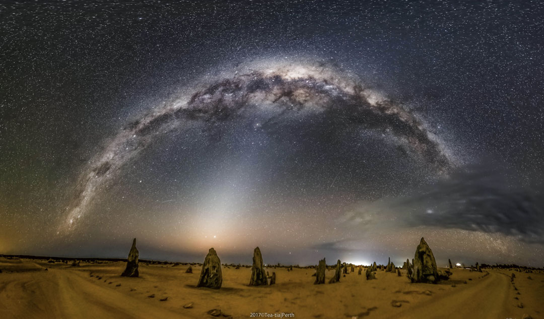 Prezentowane zdjęcie ukazuje krajobraz z wąskimi, trójkątnymi skałami pod nocnym niebem z rozpościerającym się łukiem Drogi Mlecznej.
Ziemię z niebem łączy jasne, rozmyte światło zodiakalne.
Więcej szczegółowych informacji w opisie poniżej.