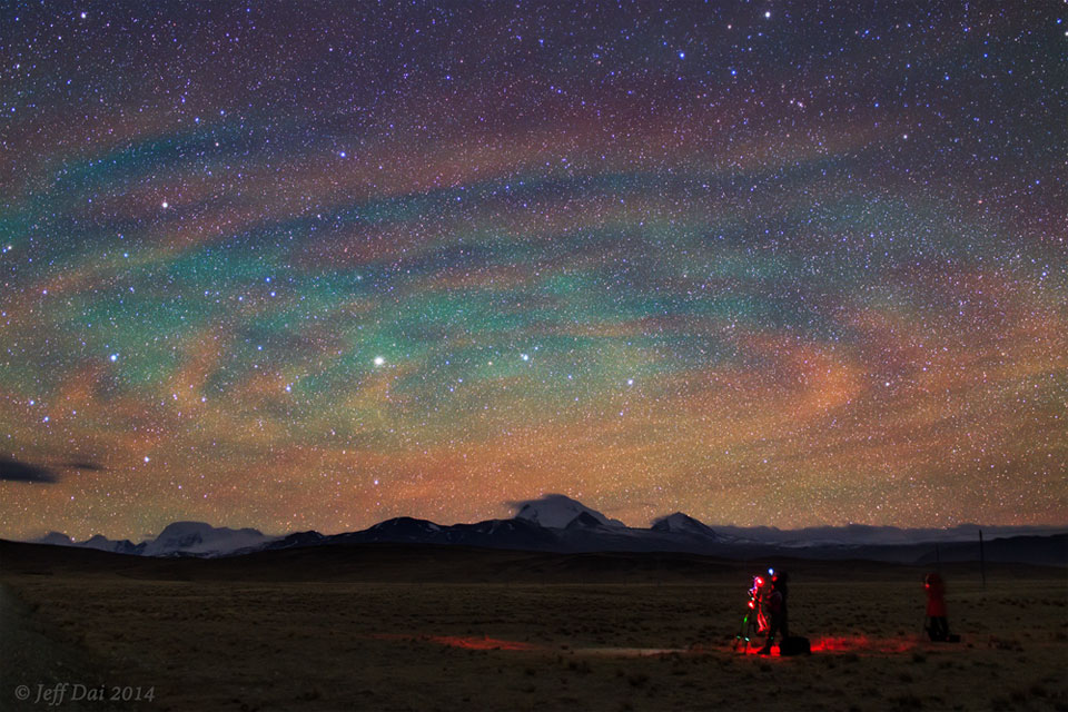 Opisywane zdjęcie pokazuje ciemne pole z oświetlonym
czerwonym światłem fotografem wykonującym zdjęcie nocnego nieba
zabarwionego zieloną poświatą i udekorowanego chmurami, które
wspólnie wyglądają jak olbrzymia spirala.
Zobacz opis. Po kliknięciu obrazka załaduje się wersja
 o&nbsp;największej dostępnej rozdzielczości.