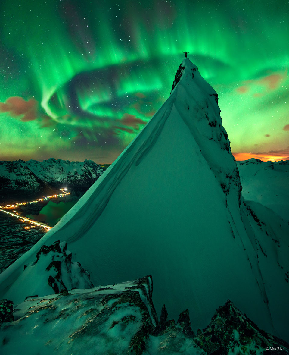 A person with the arms raised stands atop of a
rock peak covered in snow. Snow covered mountains
are all around. Green aurora swirl overhead and 
reflect off the snow. 
Więcej szczegółowych informacji w opisie poniżej.